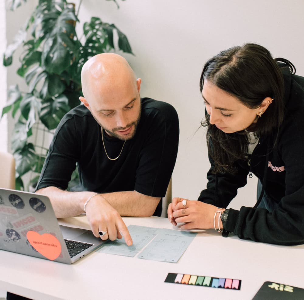 Zwei Personen arbeiten zusammen am Tisch mit Laptop und Papierplänen.