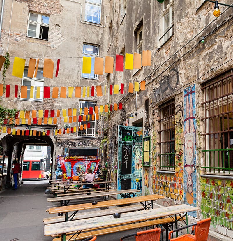 Bunte Girlanden und Graffiti in einem urbanen Hinterhof mit Bänken und Tischen.