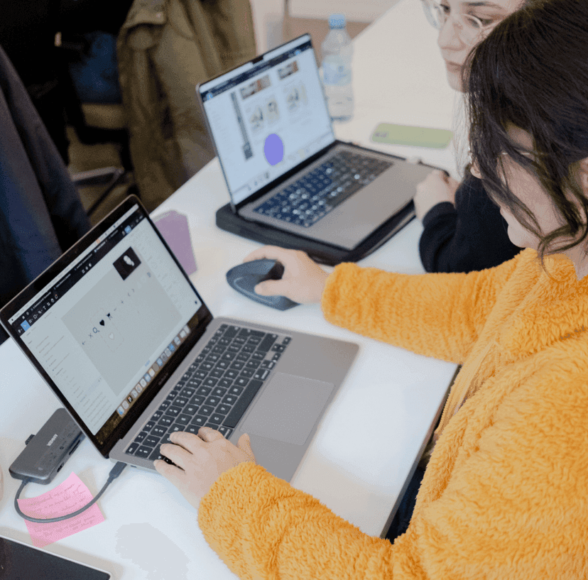 Person arbeitet an Laptop mit weiterer Person und Laptop im Hintergrund.