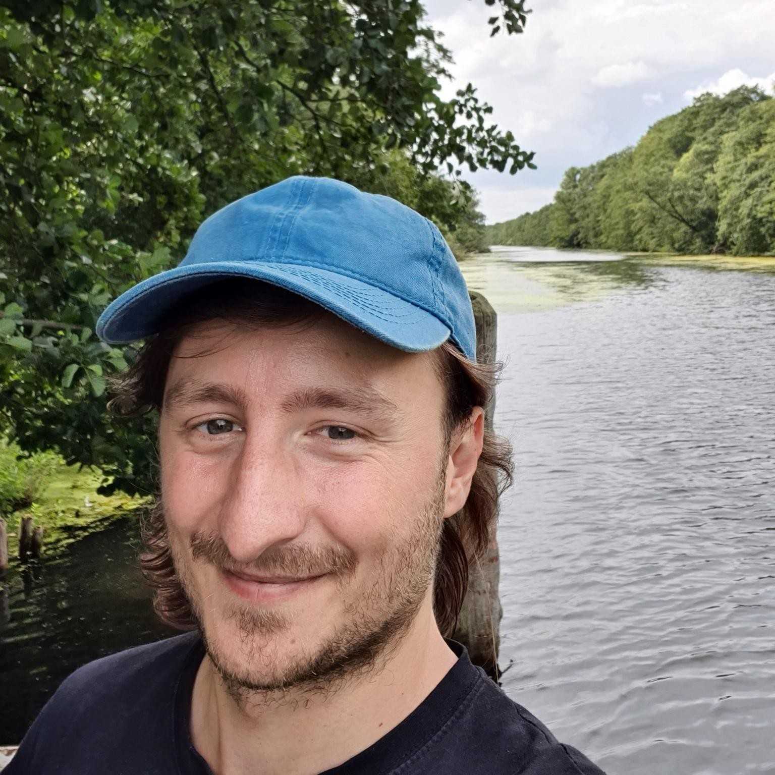Porträt eines Mannes mit blauer Kappe vor einem Fluss und Bäumen