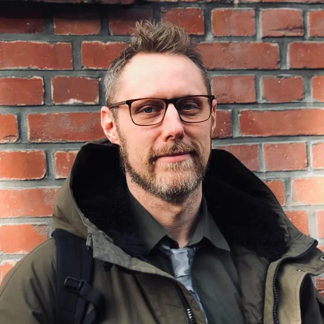 Portrait of a man with glasses in front of a red brick wall.